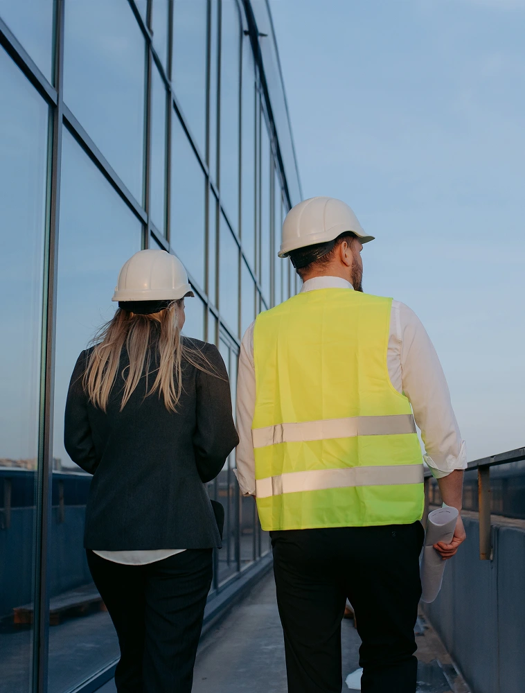 Two people in hard hats and hi-vis vests walking along glass facade